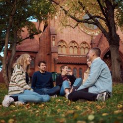 Vier junge Leute sitzen auf dem Gras vor einer Kirche und genießen die Zeit miteinander.